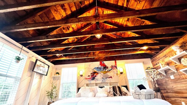 Un dormitorio con techo de madera, una cama con ropa de cama blanca y gris, un estante de pared con objetos decorativos y una ventana con persianas.