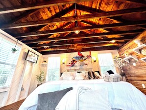 A bedroom with a wooden ceiling, a bed with white and gray bedding, a wall-mounted shelf with decorative items, and a window with blinds.