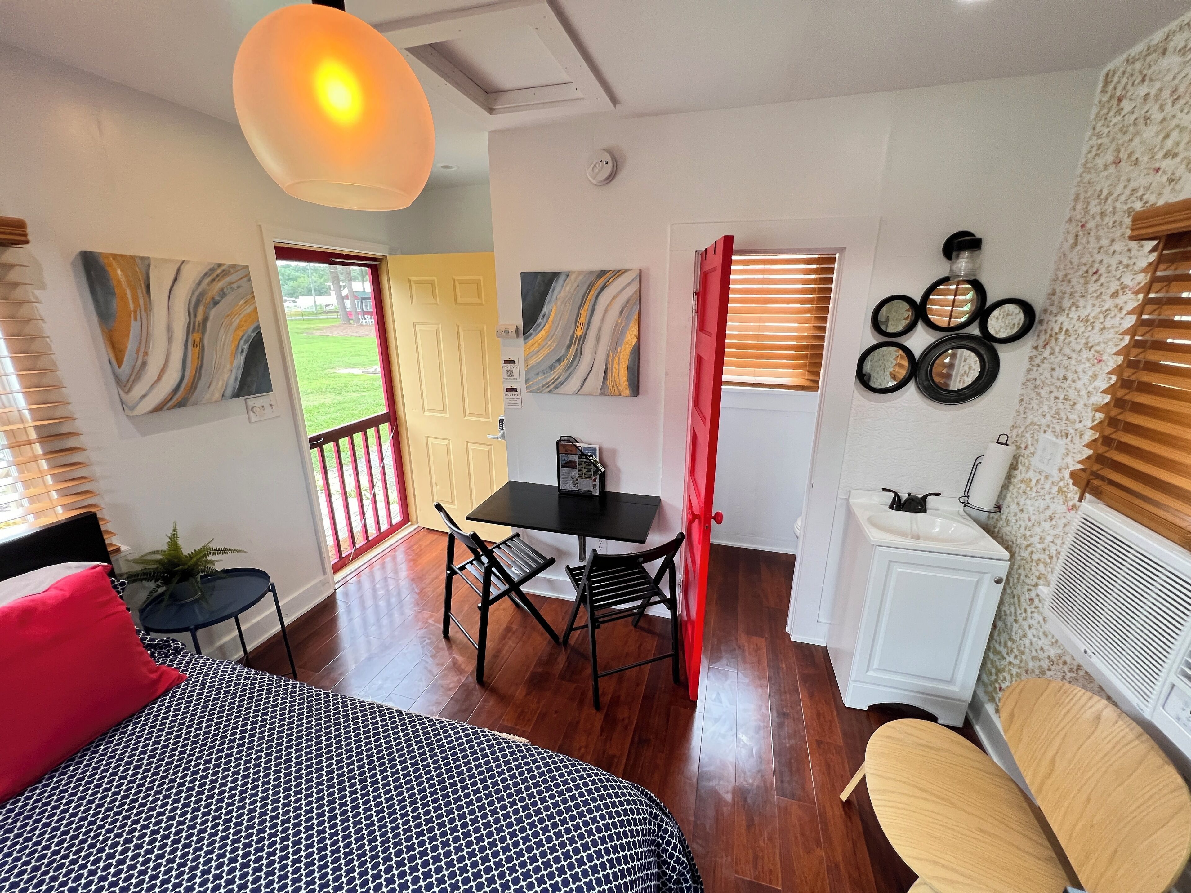 Une petite pièce comprenant un lit, une petite table et une chaise. On y trouve une porte donnant sur un balcon, ainsi qu’une fenêtre avec des stores.