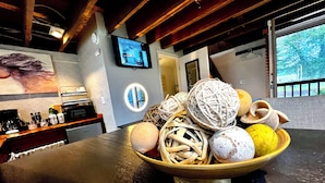 Una cocina con una mesa de madera que presenta un bol con adornos. Un televisor está montado en la pared, y una ventana ofrece vistas de la vegetación exterior.
