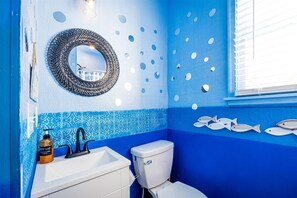 A bathroom with a blue and white color scheme, a round mirror, a toilet, a sink, and decorative fish on the wall. - Cape Charles Tiny Livin (Cape Charles)