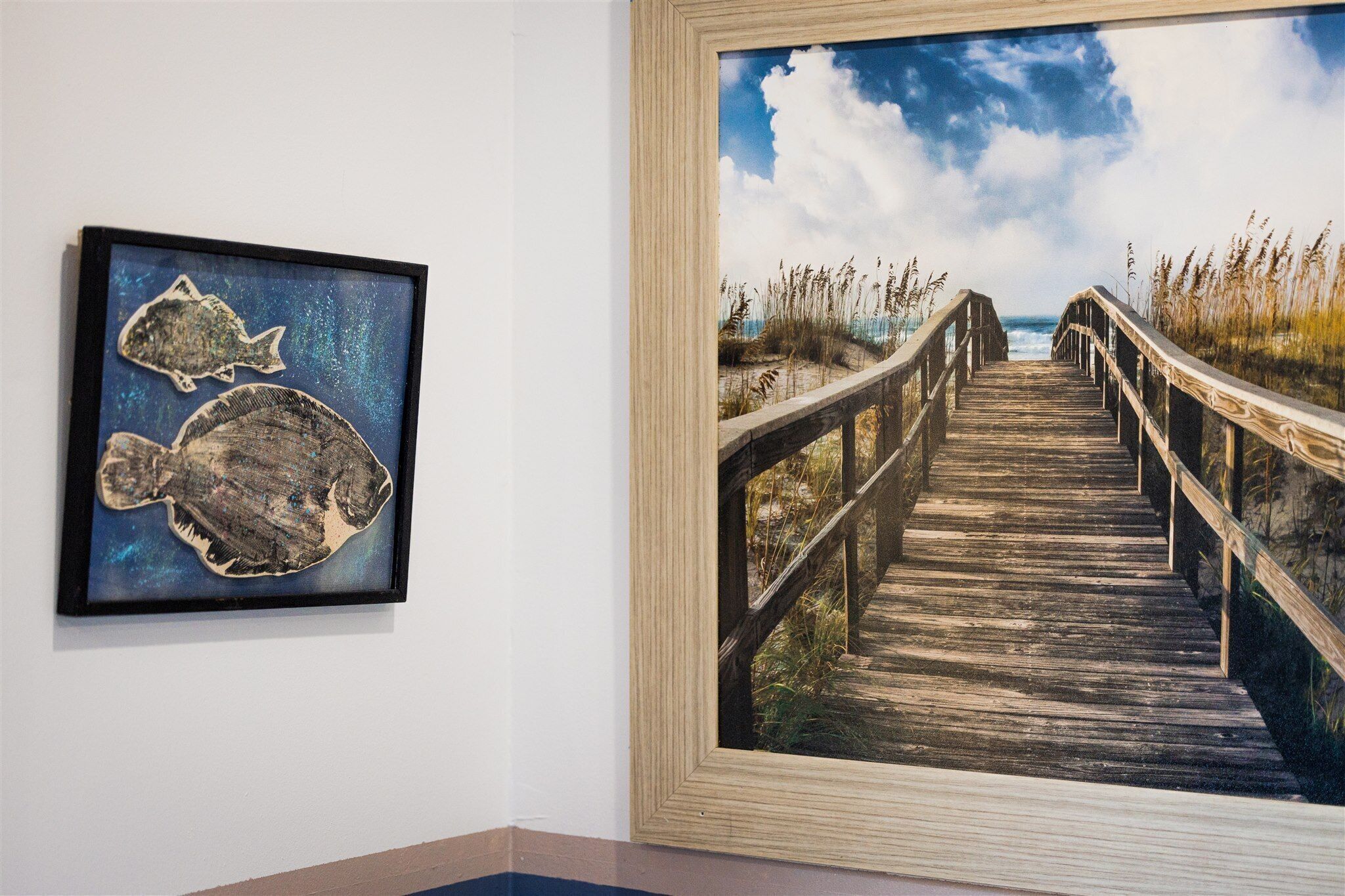 A framed artwork of two fish on the left and a wooden boardwalk leading to the sea on the right.