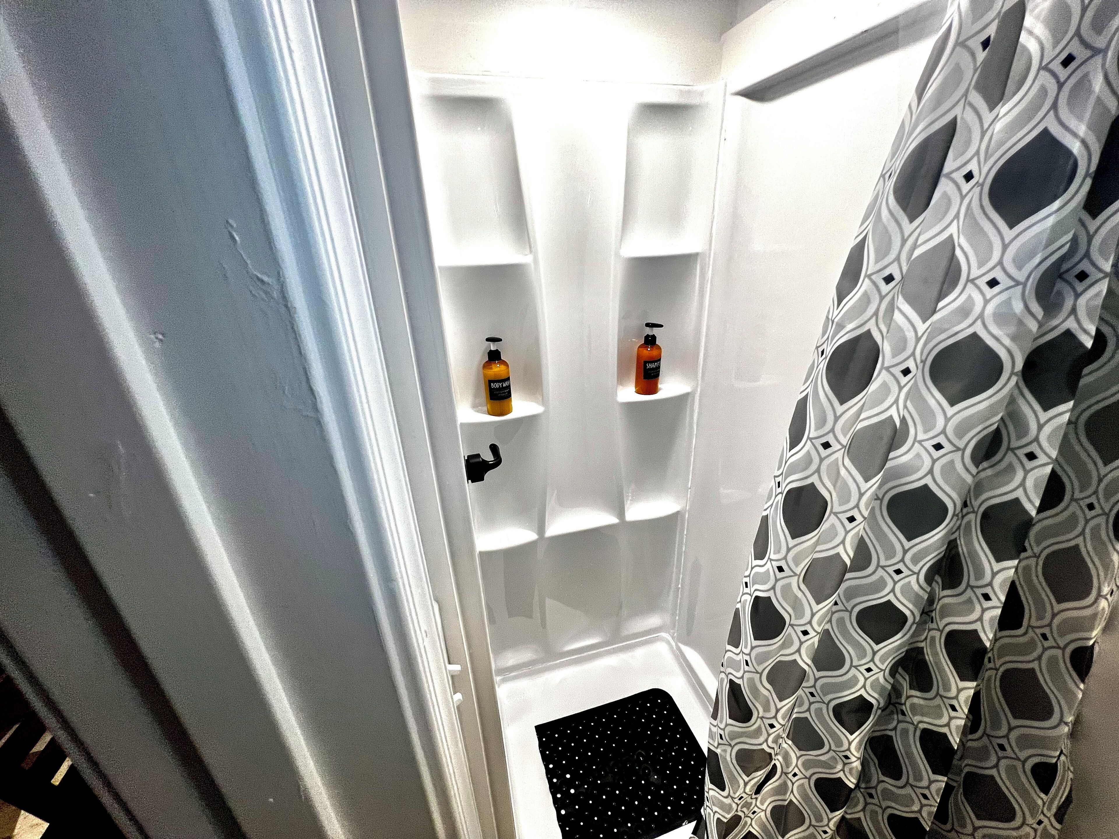 A bathroom with white walls, a shower with a geometric patterned curtain, and two shelves with bottles.