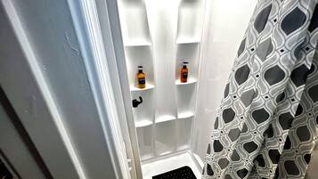 A bathroom with white walls, a shower with a geometric patterned curtain, and two shelves with bottles.