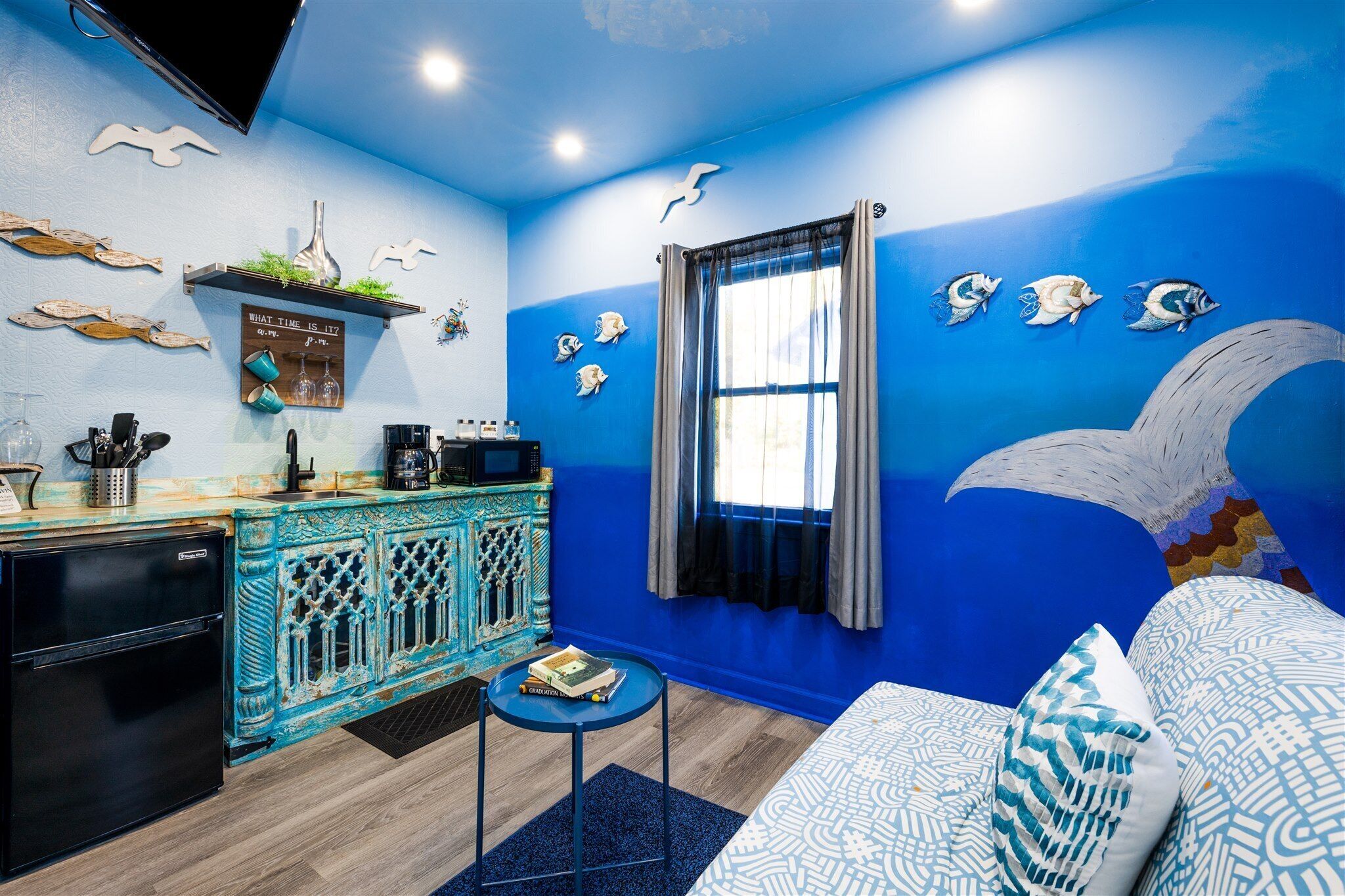 A room with a blue wall, a bed with patterned bedding, a small table with a book, a kitchenette with a black refrigerator, and decorative fish wall art.