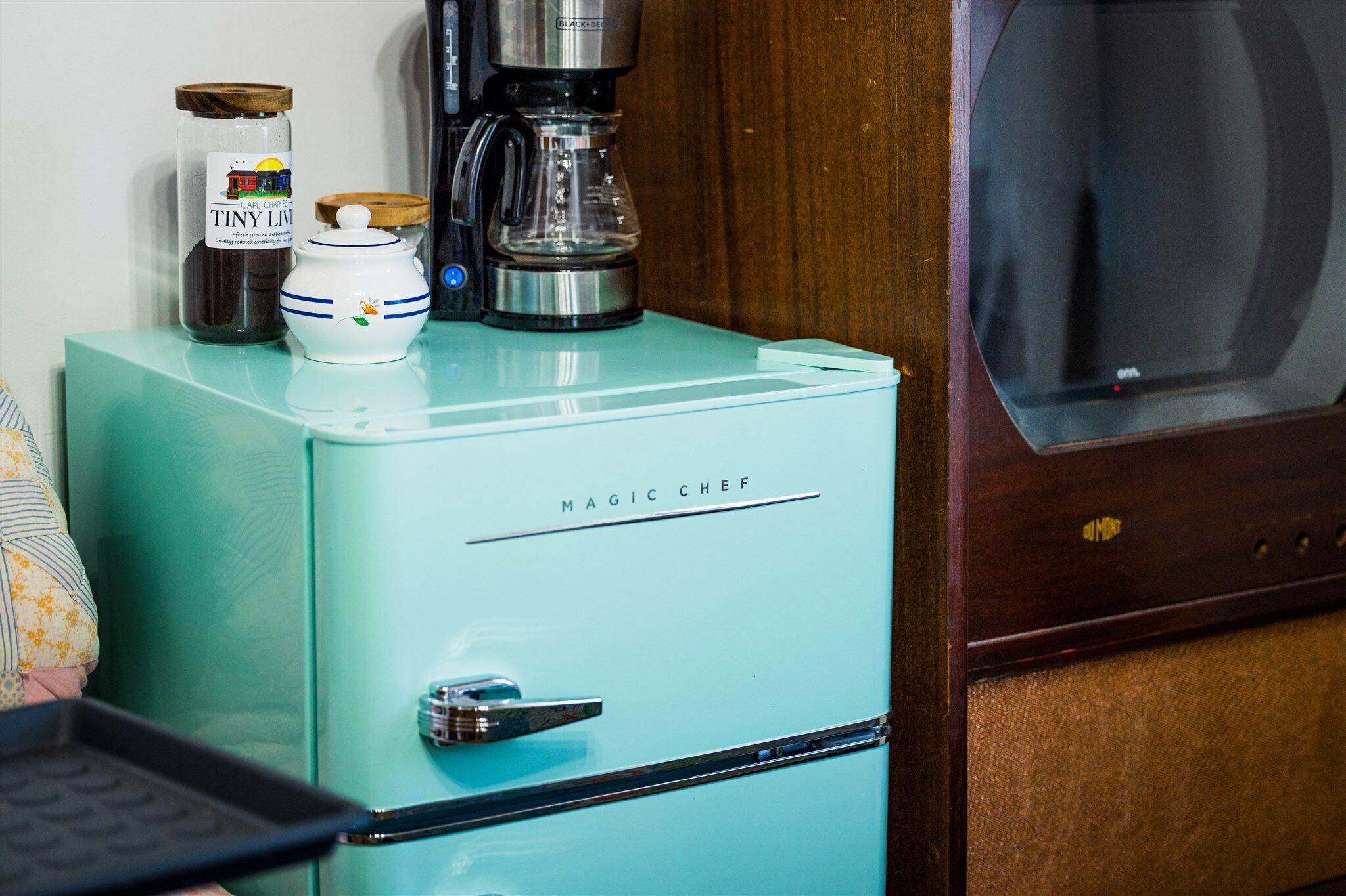 A vintage teal kitchen with a coffee maker, a jar of coffee, and a small white pot on top of a refrigerator labeled "MAGIC CHEF".