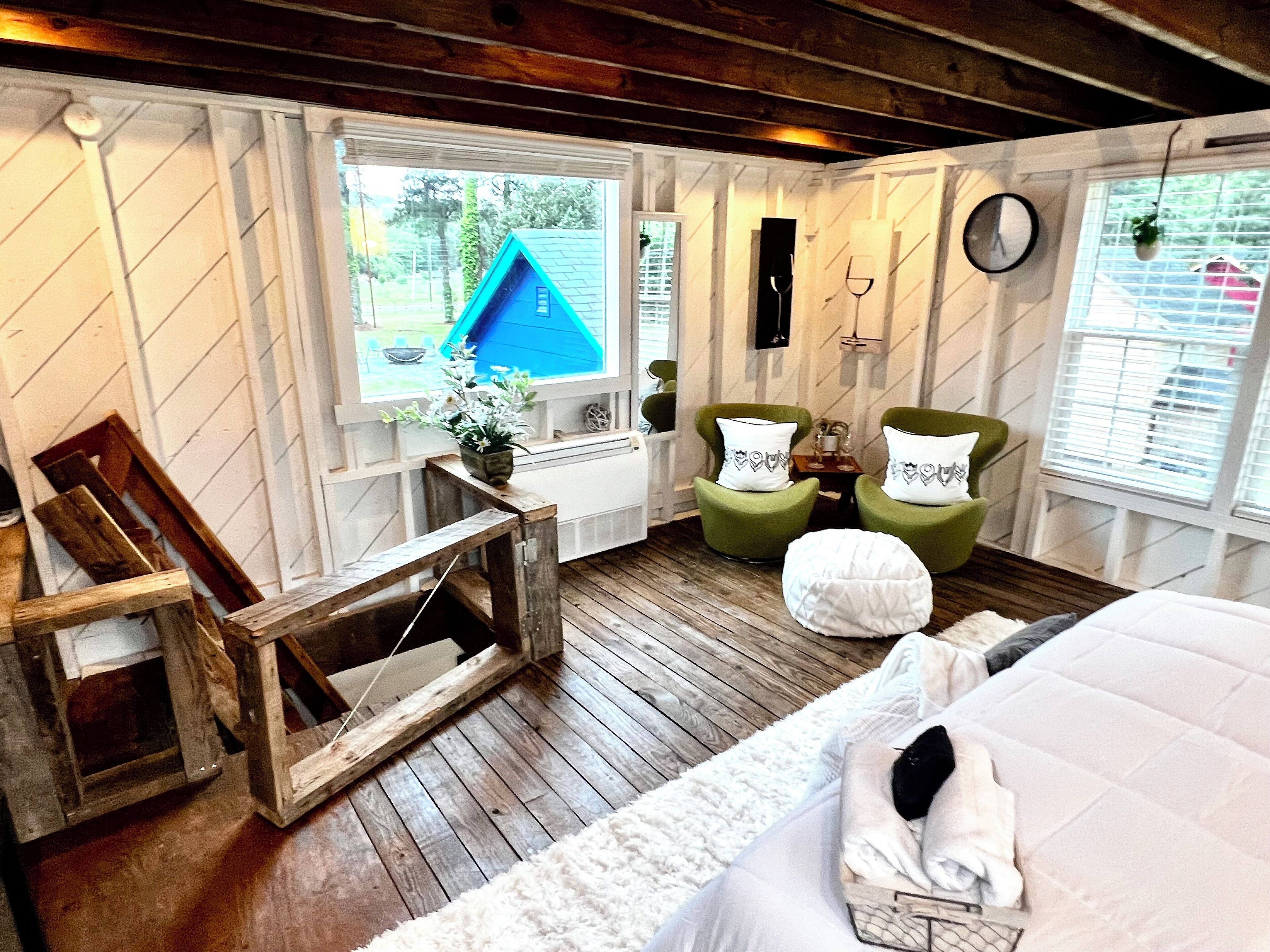 Una habitación acogedora con suelo de madera, una cama con ropa de cama blanca, dos sillones verdes, una pequeña mesa de madera y una ventana con vistas a una casa azul y vegetación.
