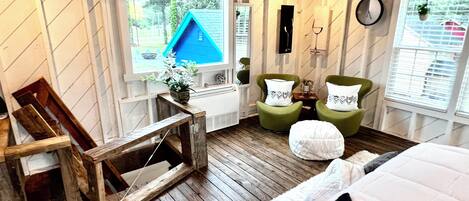 Una habitación acogedora con suelo de madera, una cama con ropa de cama blanca, dos sillones verdes, una pequeña mesa de madera y una ventana con vistas a una casa azul y vegetación.