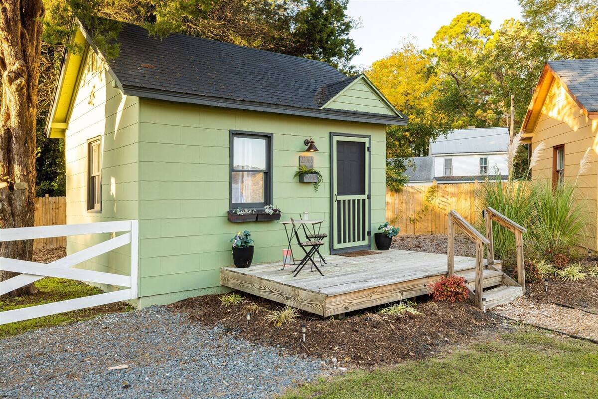 Une petite maison à un seul étage, à l’extérieur vert, avec un porche à l’avant doté d’une terrasse en bois et une clôture blanche en piquets.