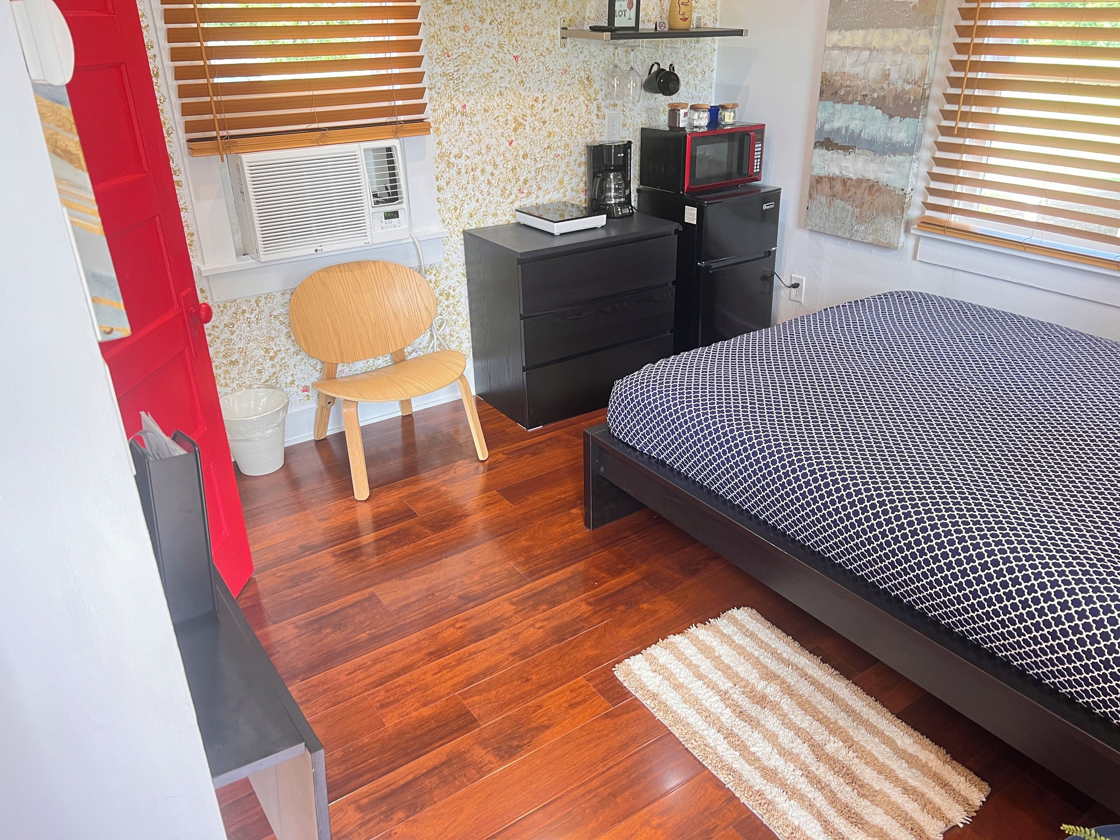 A bedroom with a bed, a chair, a dresser, a microwave, and an air conditioning unit.