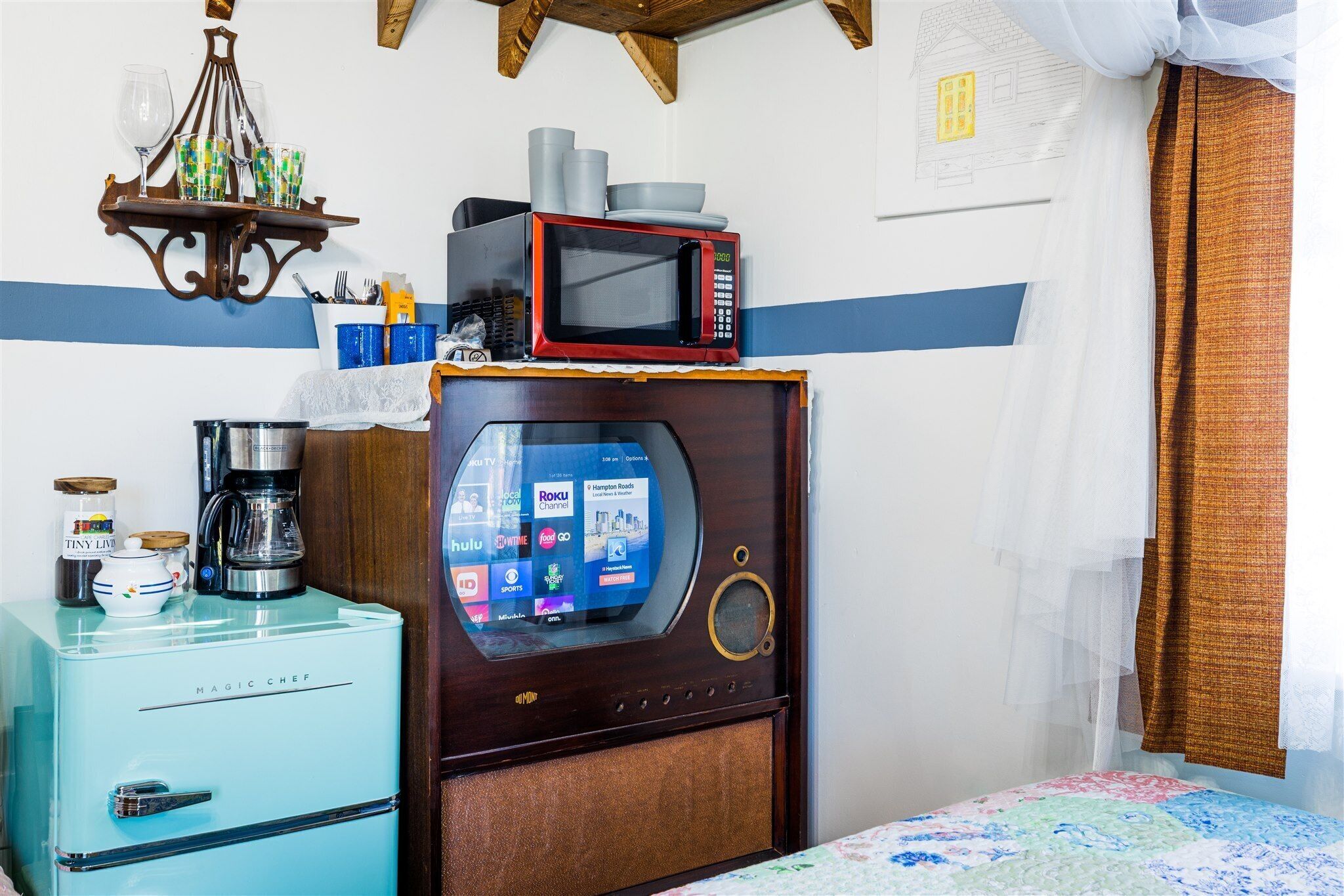 Quarto com geladeira azul, micro-ondas em cima, cafeteira e TV exibindo diversos canais.