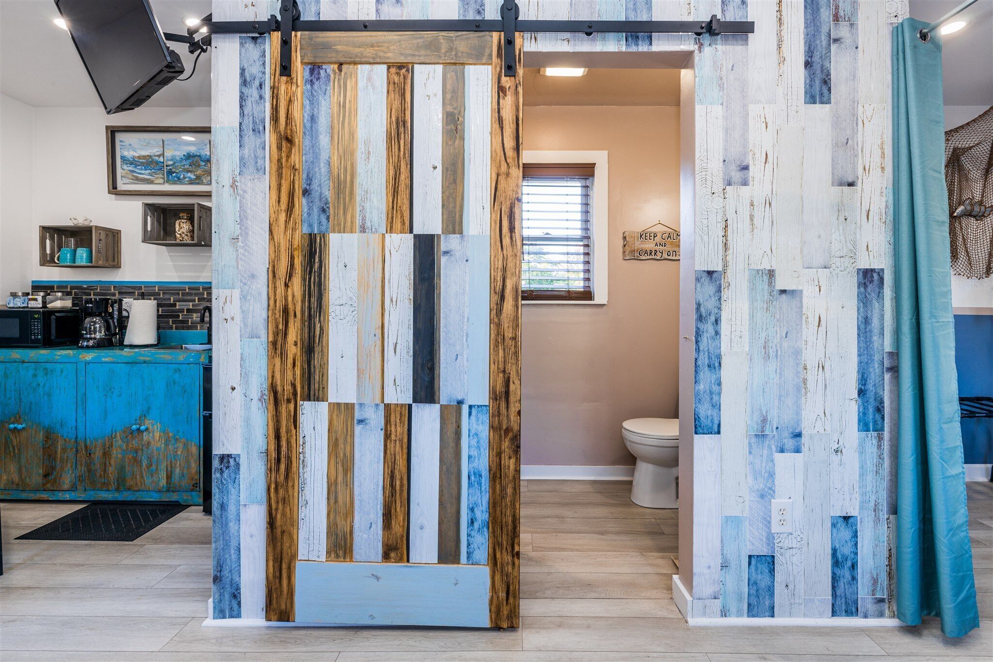 A bathroom with a rustic wooden door, a blue shower curtain, and a sign that reads "KEEP CALM AND CARRY ON."