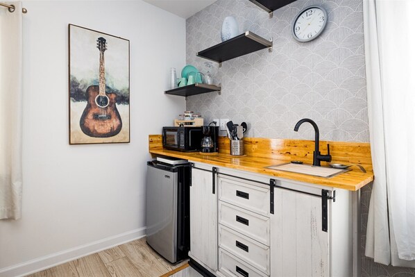 A compact kitchen with a wooden countertop, a sink, a microwave, and a refrigerator. There are two cabinets with black handles and a clock on the wall. - Cape Charles Tiny Livin (Cape Charles)