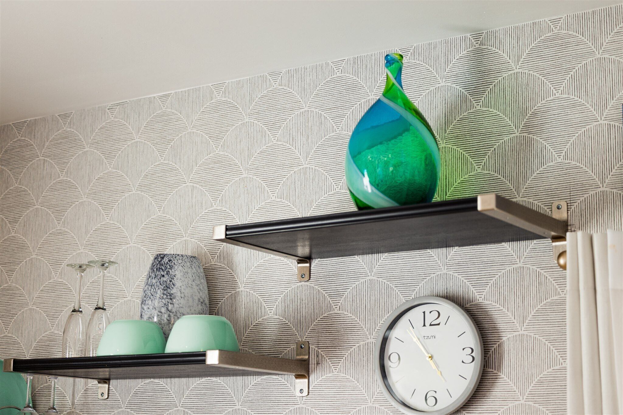 Un mur recouvert d’un papier peint à motifs géométriques, un vase vert, une horloge et des étagères ornées d’objets décoratifs.