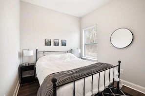 A bedroom with a bed, a nightstand, a lamp, a window with blinds, and three framed pictures on the wall.