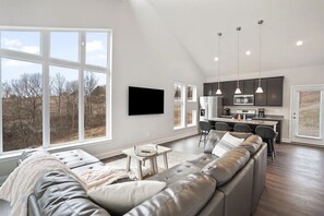 Una sala de estar moderna con un amplio sofá seccional gris, un televisor de pantalla plana montado en la pared y una cocina con armarios oscuros e iluminación colgante.