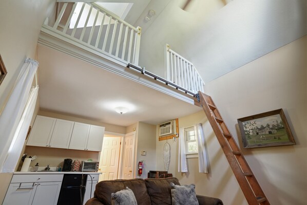 A loft bedroom with a sloped ceiling, a wooden ladder, a kitchenette, and a dining area.