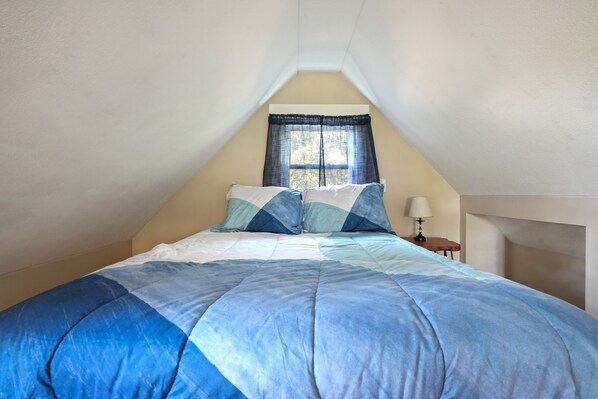 Une chambre avec un plafond en pente, un lit recouvert d’une couette bleue et blanche, une fenêtre avec des rideaux bleus et une table de chevet avec une lampe.