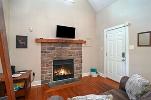 A living room with a stone fireplace, a mounted TV, a door, and a wooden floor. - Love Shack Cabin | Hocking Hills, Couples Retreat (McArthur)