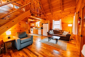 A wooden cabin interior with a staircase, a living room with a sofa and coffee table, and a kitchen area.