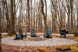 Un camping arbolado con tres sillas Adirondack, un brasero y una parrilla.