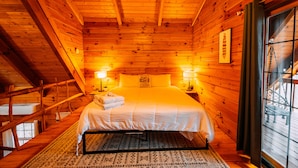 A wooden cabin bedroom with a bed, two bedside lamps, a window with a view, and a cabin sign on the wall. - Oak Cabin | Hocking Hills, Romantic Hot Tub (New Plymouth)