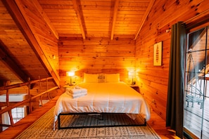 Une chambre aménagée dans une cabane en bois, avec un lit, deux lampes de chevet, une fenêtre donnant sur un paysage agréable et une enseigne indiquant qu’il s’agit d’une cabane, fixée au mur.
