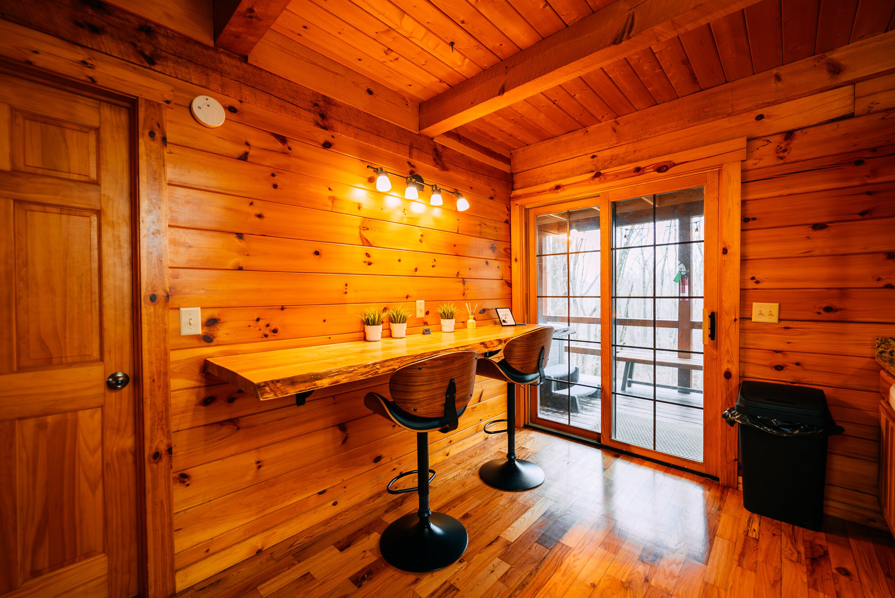 A wooden interior with a bar area, two bar stools, a trash can, and a door leading outside.