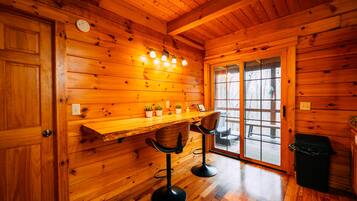 A wooden interior with a bar area, two bar stools, a trash can, and a door leading outside.