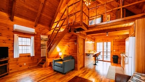 A log cabin interior with a staircase, a sofa, a television, and a kitchen area. - Oak Cabin | Hocking Hills, Romantic Hot Tub (New Plymouth)