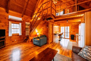 A log cabin interior with a staircase, a sofa, a television, and a kitchen area.