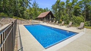 Une piscine rectangulaire dotée d’une terrasse en bois et de chaises longues, entourée d’une clôture en métal, avec une petite structure ressemblant à un chalet en arrière-plan.