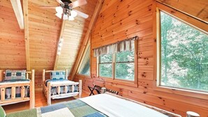 A cozy cabin room with a sloped ceiling, two wooden beds with plaid bedding, a large window with curtains, and a ceiling fan. - Grizzly Ridge | Hocking Hills, Pool, Hot Tub (Logan)