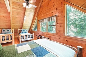A cozy cabin room with a sloped ceiling, two wooden beds with plaid bedding, a large window with curtains, and a ceiling fan.