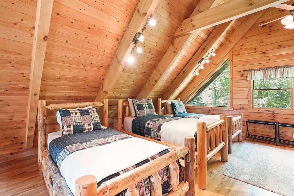 A cozy cabin room with two wooden beds, a large window, and a ceiling fan.