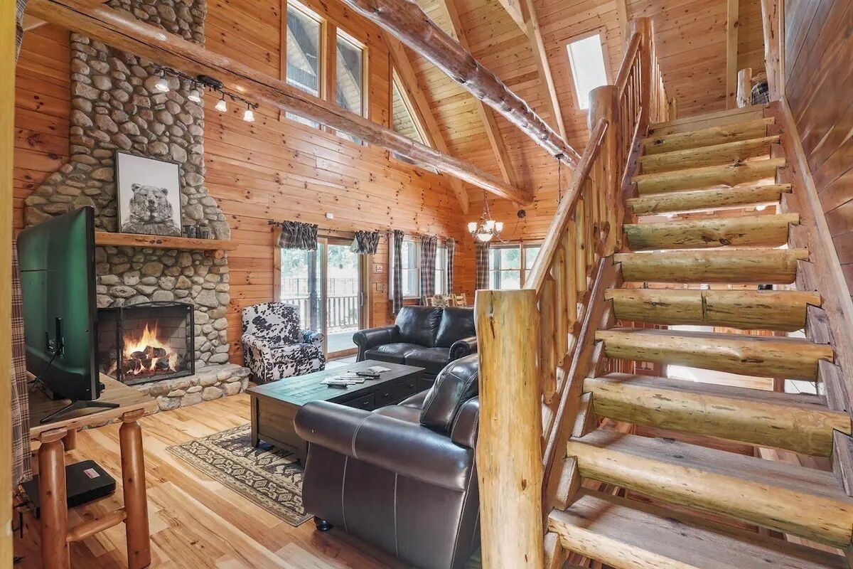 Ein rustikales Blockhaus-Interieur mit Kamin aus Stein, Ledersofa, einer Holztreppe und einem Fernseher.