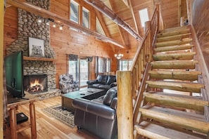 A log cabin interior with a stone fireplace, leather sofa, wooden staircase, and a television.