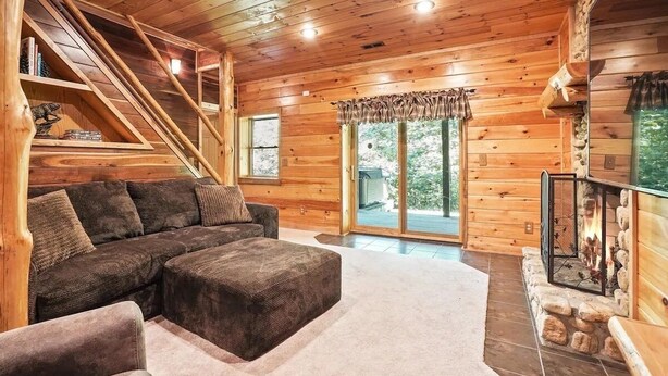 Un intérieur chaleureux de chalet en bois, avec un canapé d’angle, une cheminée, un escalier et une grande fenêtre.