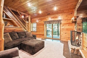 A cozy log cabin interior with a sectional sofa, a fireplace, a staircase, and a large window. - Grizzly Ridge | Hocking Hills, Pool, Hot Tub (Logan)