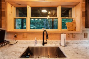 Una cocina con pileta de acero inoxidable, armarios de madera, un ventanal con vista a árboles y una encimera con una botella de jabón de manos.