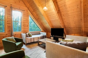 Une charmante cabane en bois avec une chambre à l’étage, un espace de vie comprenant un canapé et des fauteuils, ainsi qu’une télévision.