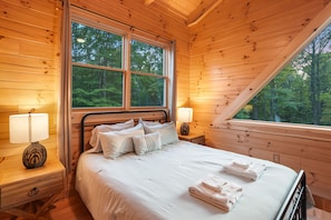 A wooden cabin bedroom with a large window, a bed with pillows, and two bedside lamps.