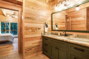 Een houten cabin met een bed, een badkamer met een wastafel en spiegel, en uitzicht op de natuur door een raam.