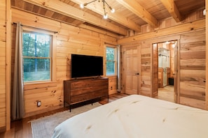 A wooden cabin bedroom with a large bed, a TV on a dresser, and a window overlooking the forest.