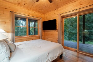 A wooden cabin bedroom with a bed, a TV mounted on the wall, and large windows offering a view of the forest.