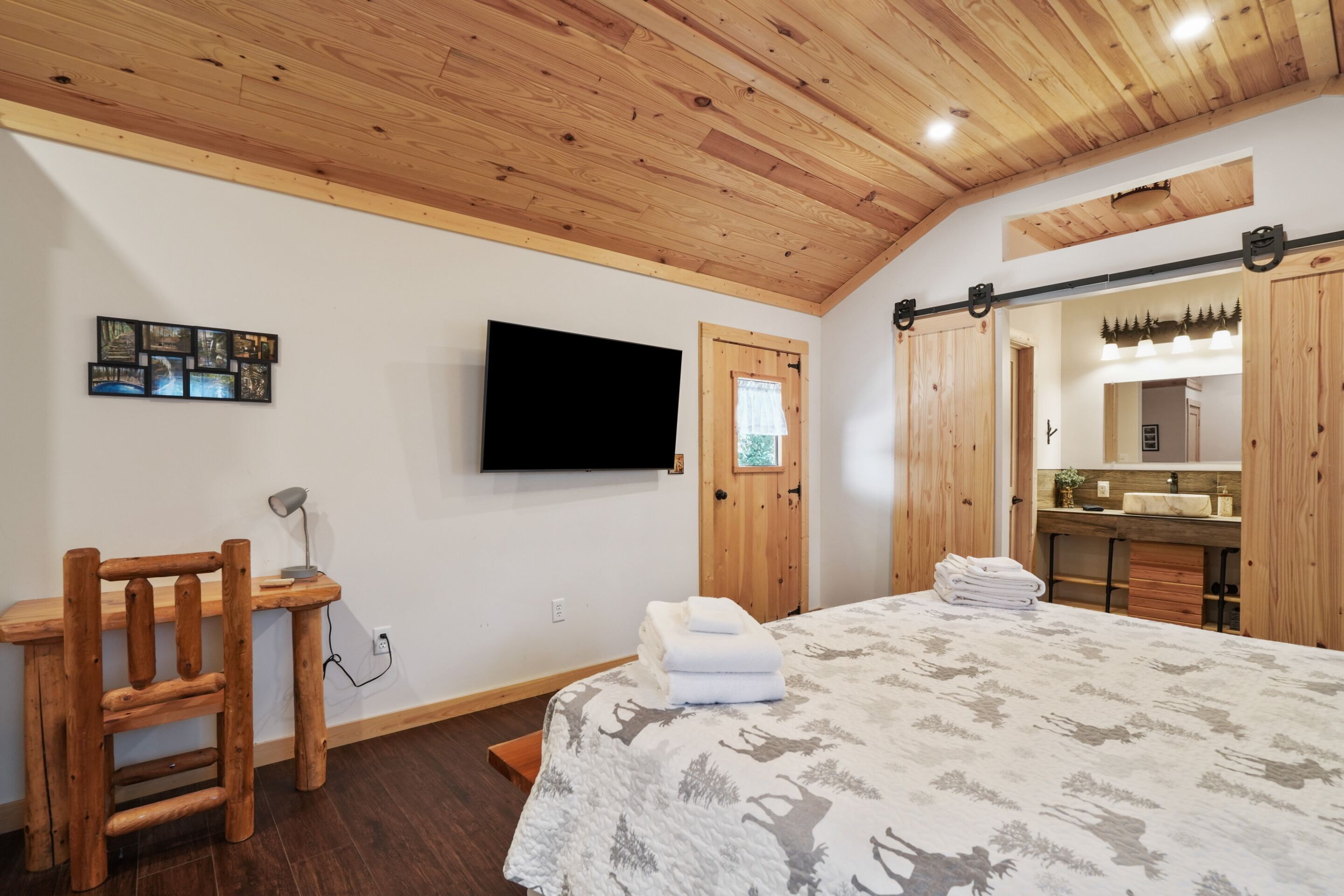 A bedroom with a wooden ceiling, a bed with a patterned bedspread, a desk with a chair, a TV mounted on the wall, and a bathroom visible through a door.