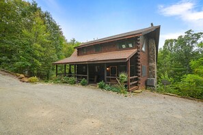 A two-story log cabin with a covered porch, surrounded by dense forest. - Hidden Haven | Hocking Hills, Sleeps 8, Hot Tub (Laurelville)