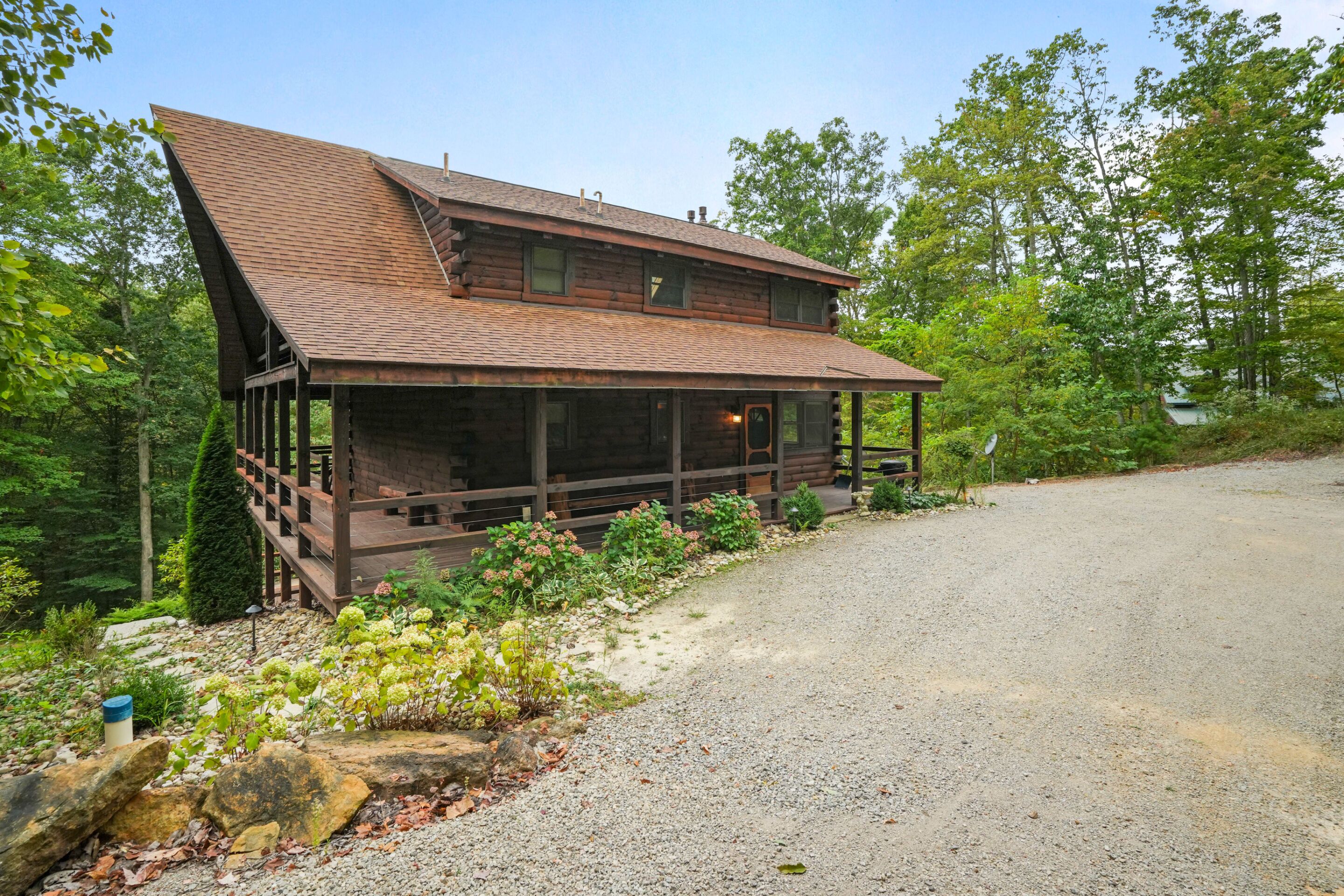 A rustic log cabin with a steeply pitched roof, a porch with wooden railings, and a gravel driveway.