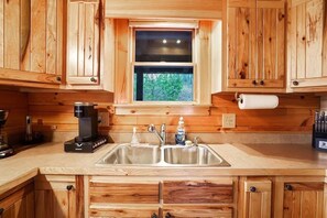 A wooden kitchen with a double sink, a coffee maker, and a window with a view of trees. - Hidden Haven | Hocking Hills, Sleeps 8, Hot Tub (Laurelville)