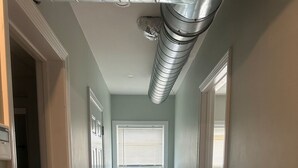 A hallway with a light green wall, wooden flooring, and a window with blinds. - Almira Properties Unit 3 (Providence)